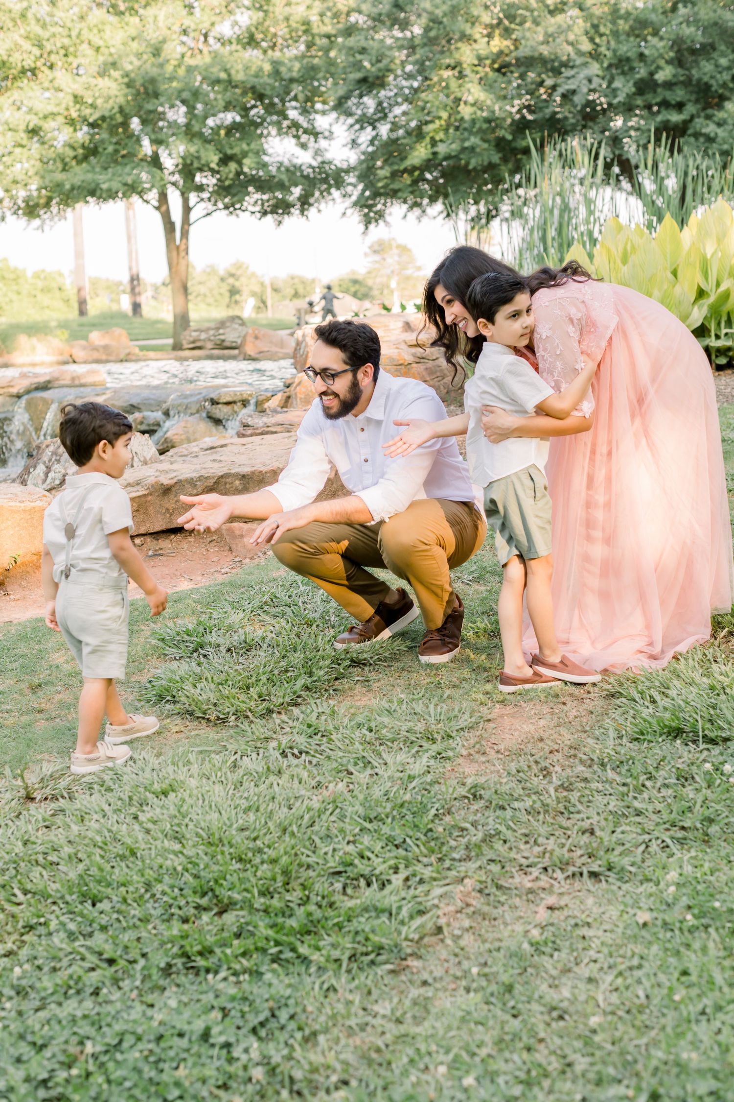 A sweet family moment captured outdoors as they interact playfully on grass near rocks and trees.