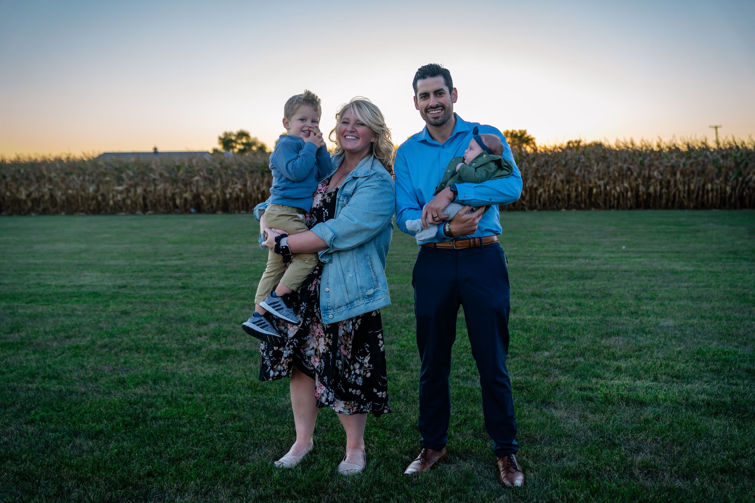 A family stands together at sunset in front of a cornfield on a grassy field.