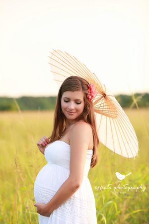 Maternity photo shoot featuring woman in white dress holding parasol in sunlit field.