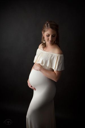 Elegant maternity portrait against dark background with off-shoulder cream dress.