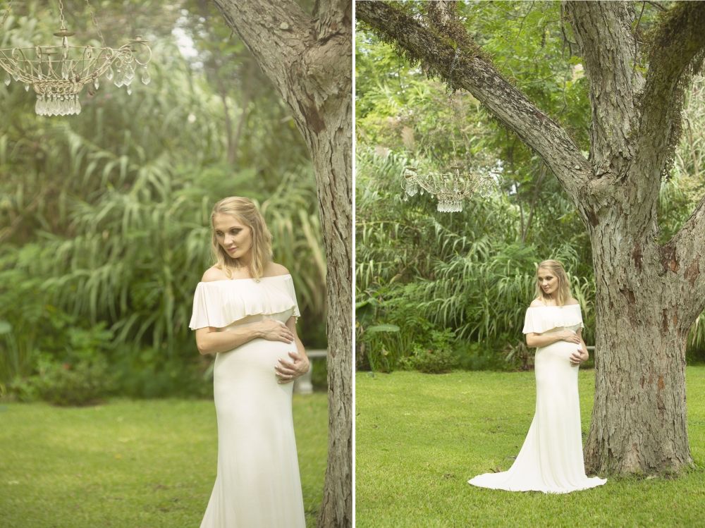 A maternity photo shoot in a garden setting with a white off-shoulder dress beside a large tree.