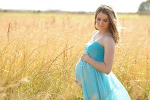 Expecting mother in flowing turquoise dress poses in golden wheat field at sunset.