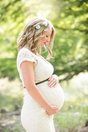 Romantic maternity portrait in white lace dress with floral headpiece outdoors.