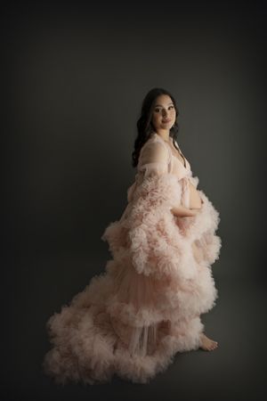 Dramatic maternity portrait in flowing pink tulle gown against dark background.