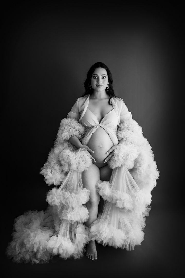 Artistic maternity photo in black and white featuring a flowing tulle robe with dramatic ruffles against dark background.