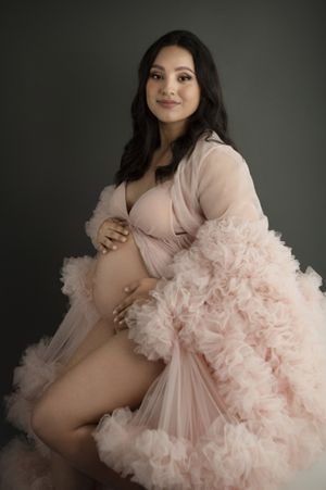 Maternity portrait in soft pink featuring romantic ruffled robe against dark backdrop.