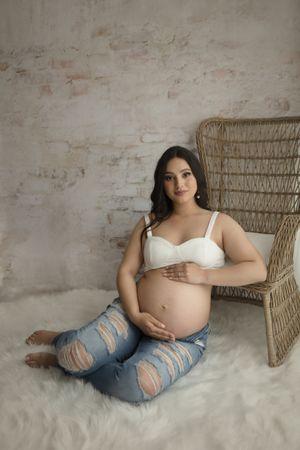 Pregnancy photoshoot with woman in white crop top and distressed jeans sitting beside rattan chair.