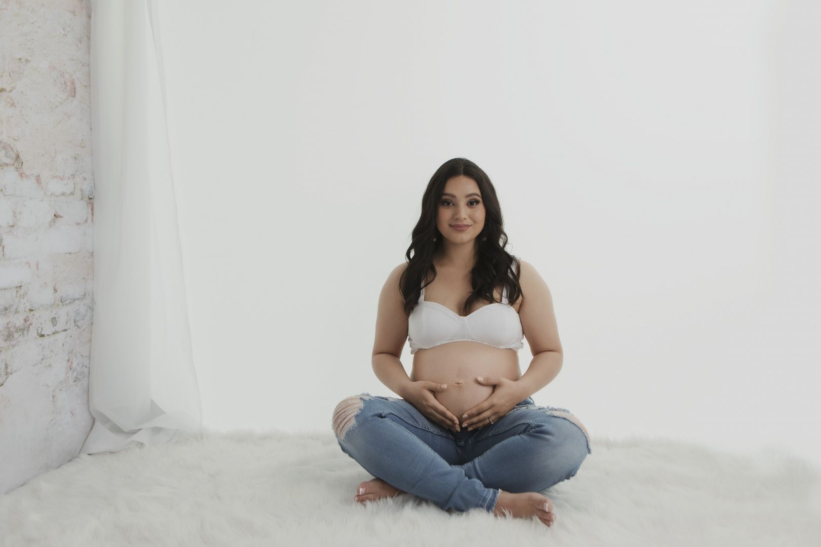Minimalist maternity portrait with expecting mother in cross-legged pose against white background.