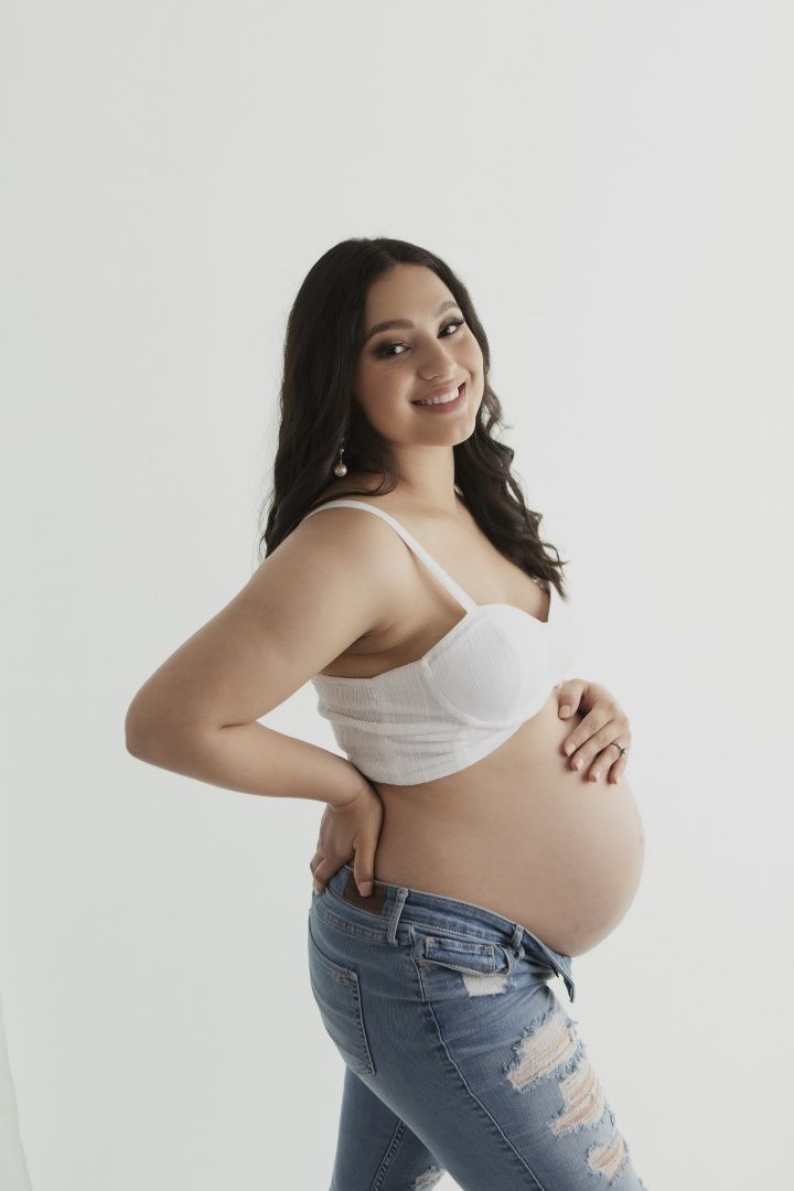 Joyful maternity portrait against white background featuring distressed jeans and white crop top.