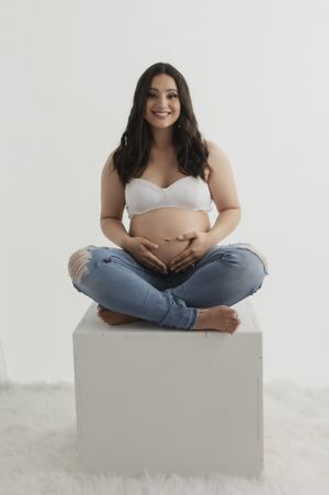 Expectant mother in casual attire sits cross-legged on white cube against plain background.