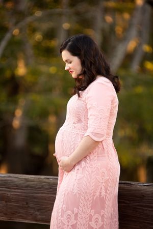 Pregnant woman in pink lace dress poses against autumn trees.