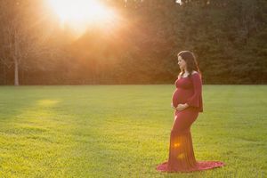Silhouette of pregnant woman in red dress standing in sunlit field.