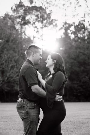 Black and white maternity photo of couple embracing outdoors at sunset.