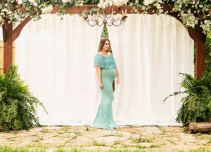 Woman in turquoise gown poses under floral arch with white backdrop.