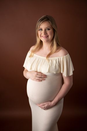 Maternity portrait with flowy cream off-shoulder dress against brown backdrop.