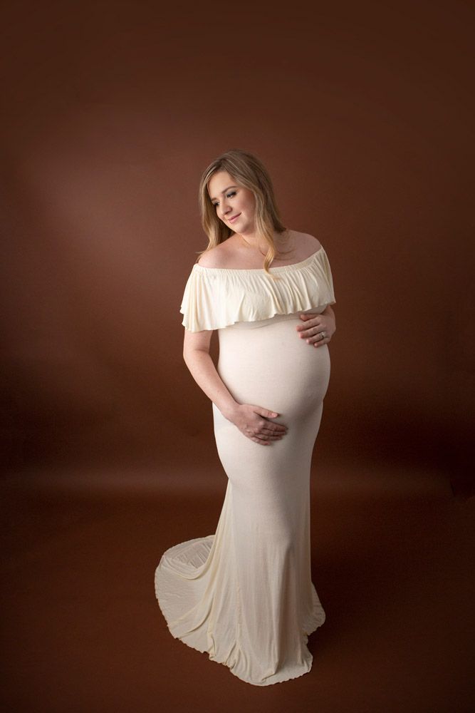 Full length maternity portrait in cream flowing gown against warm brown background.