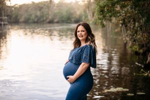 Expectant mother in denim dress poses by lake at golden hour.