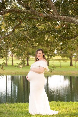 An expectant mother in a flowing white off-shoulder gown poses elegantly by a tranquil pond under tree branches.