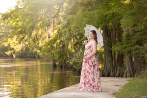 Maternity photo of woman in floral pink dress standing beside a scenic waterway during golden hour.