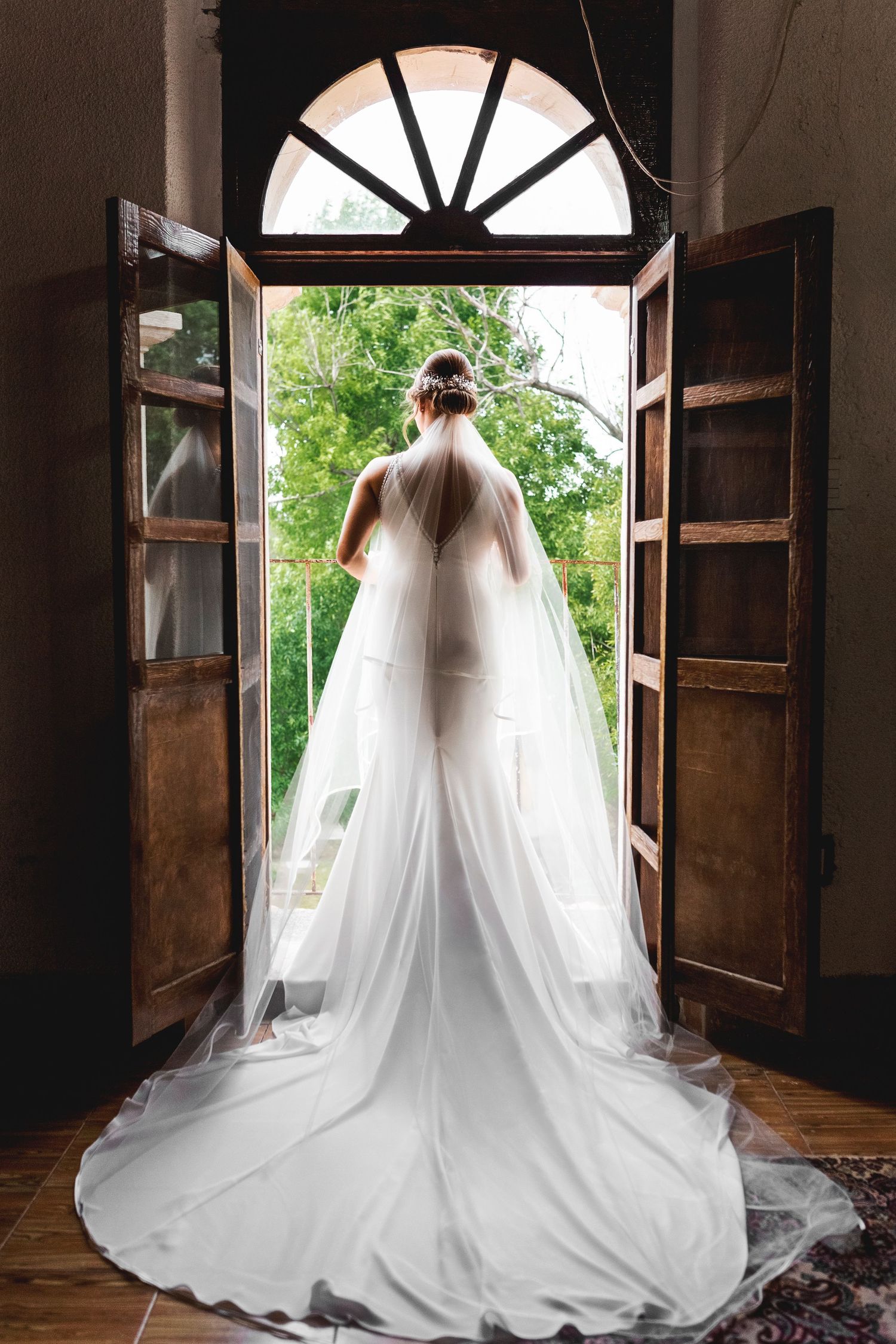 A flowing white wedding dress and veil photographed from behind in an elegant doorway with arched window.