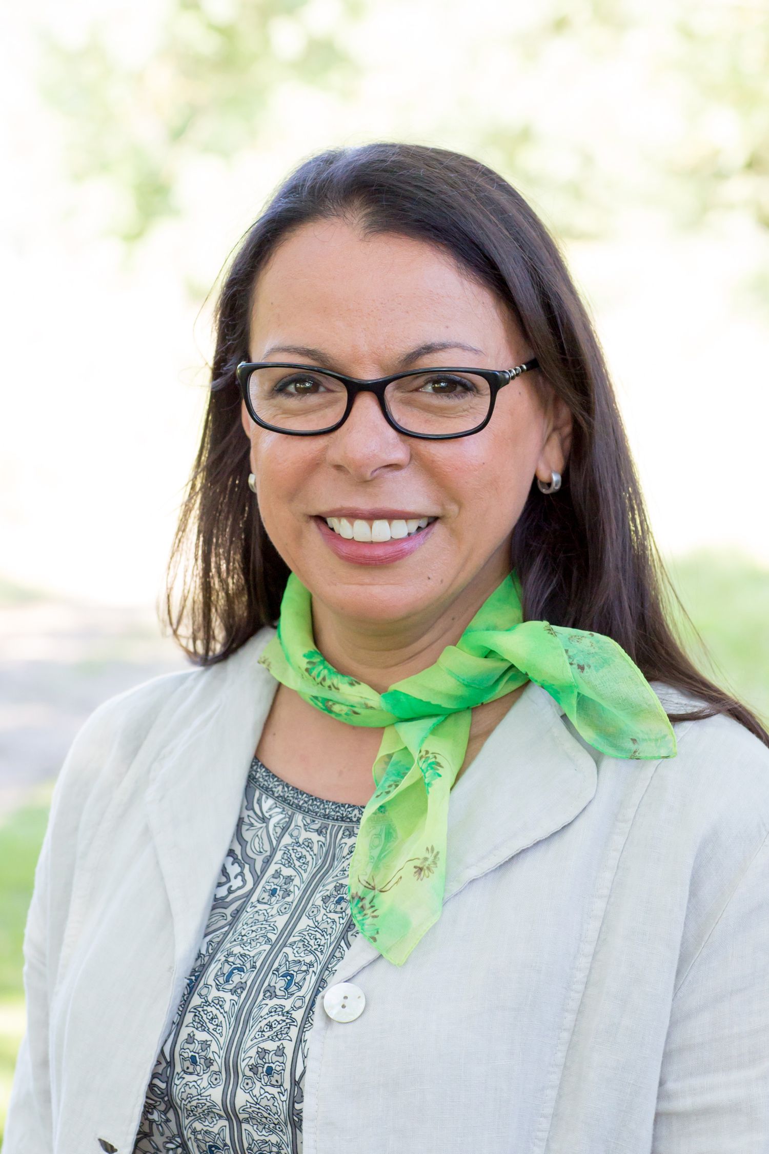 Professional headshot of a person wearing glasses, a light blazer and green scarf against a blurred background during business branding pho.