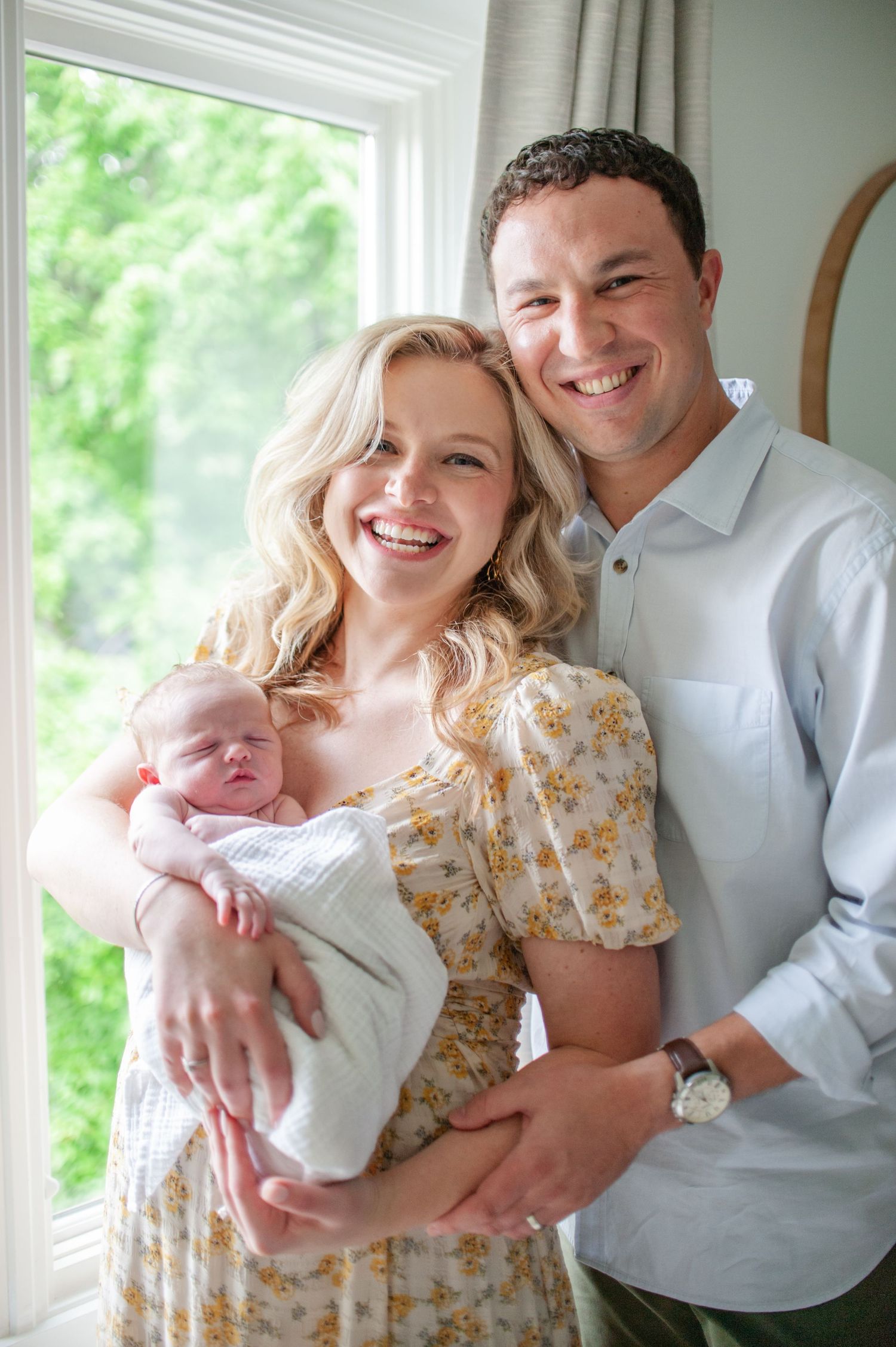 mom and dad close together in front of a window and smiling at the camera, mom is holding baby boy in her arms