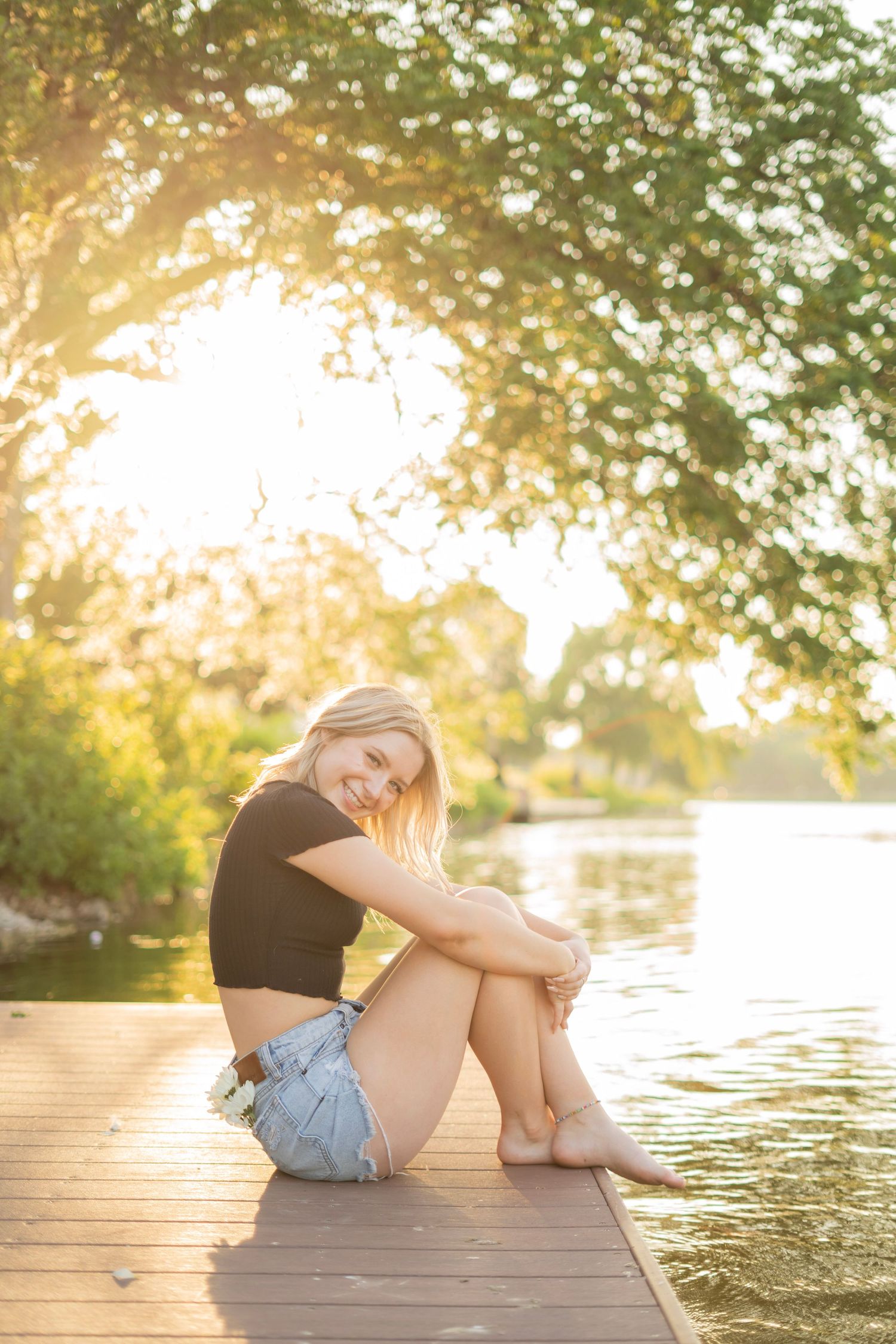 A blonde woman in a black crop top and denim shorts sits on a wooden dock by a sunlit lake.