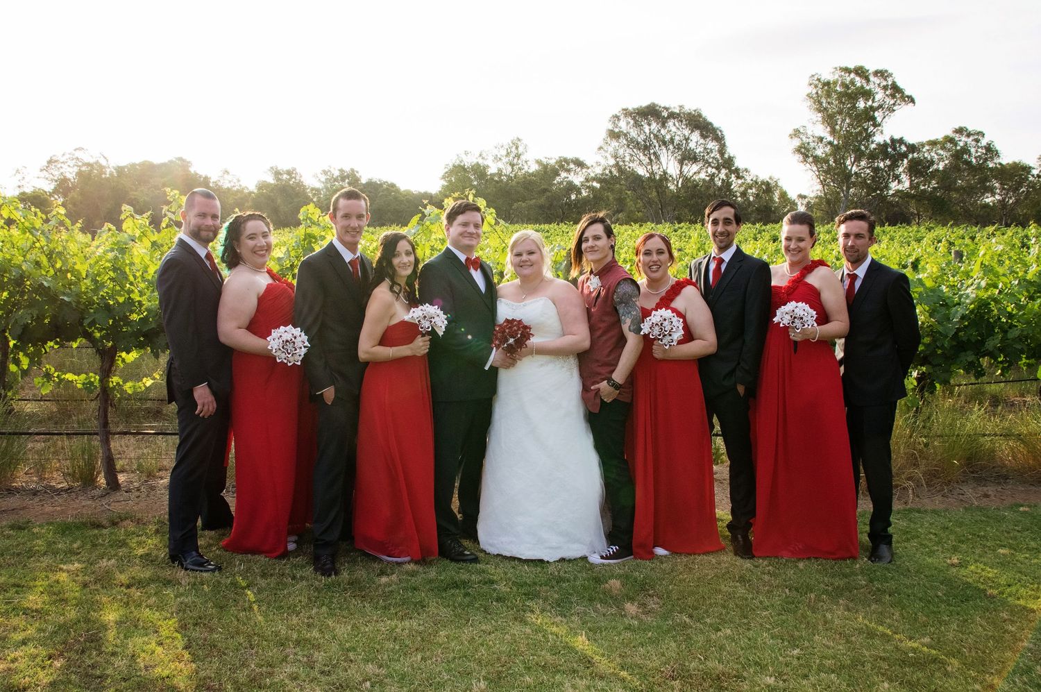 bridal party in red and black posing with bride and groom in wineyard