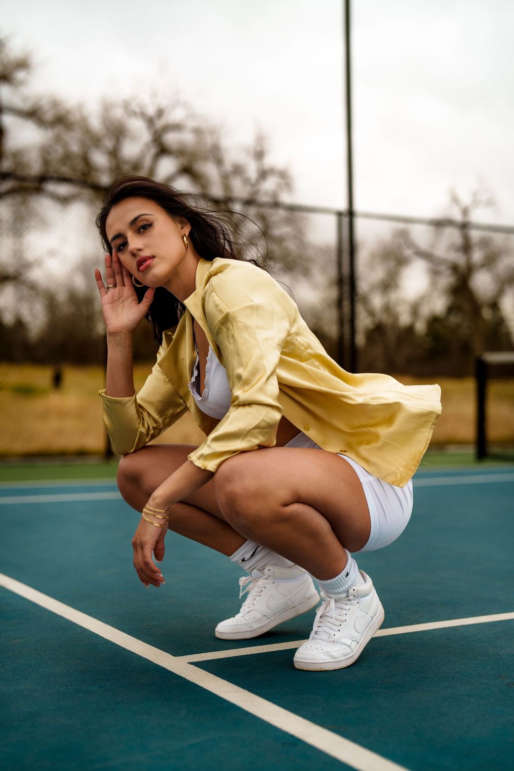 Athletic poses on a tennis court wearing a yellow top and white sneakers.
