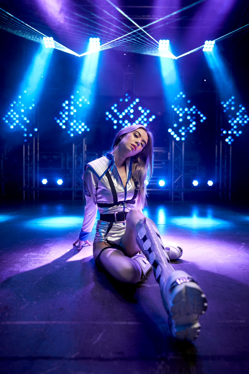 Performer in stylish outfit posing on stage with dramatic blue lighting and LED displays.