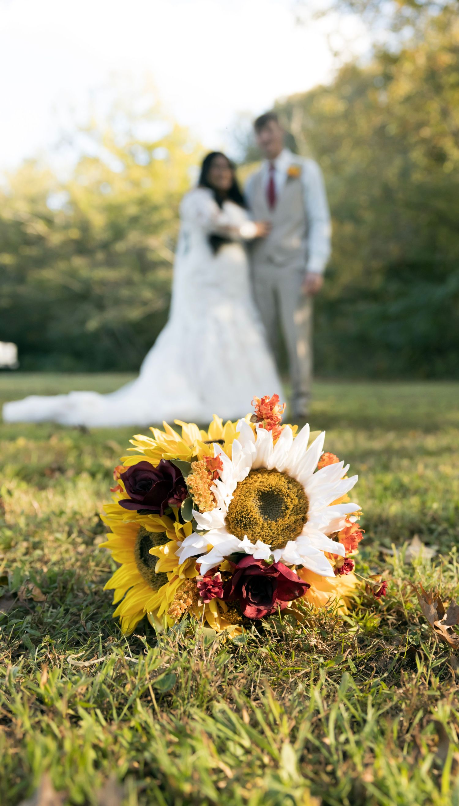 A sunflower and rose bouquet in focus with a blurred newlywed couple in the background.