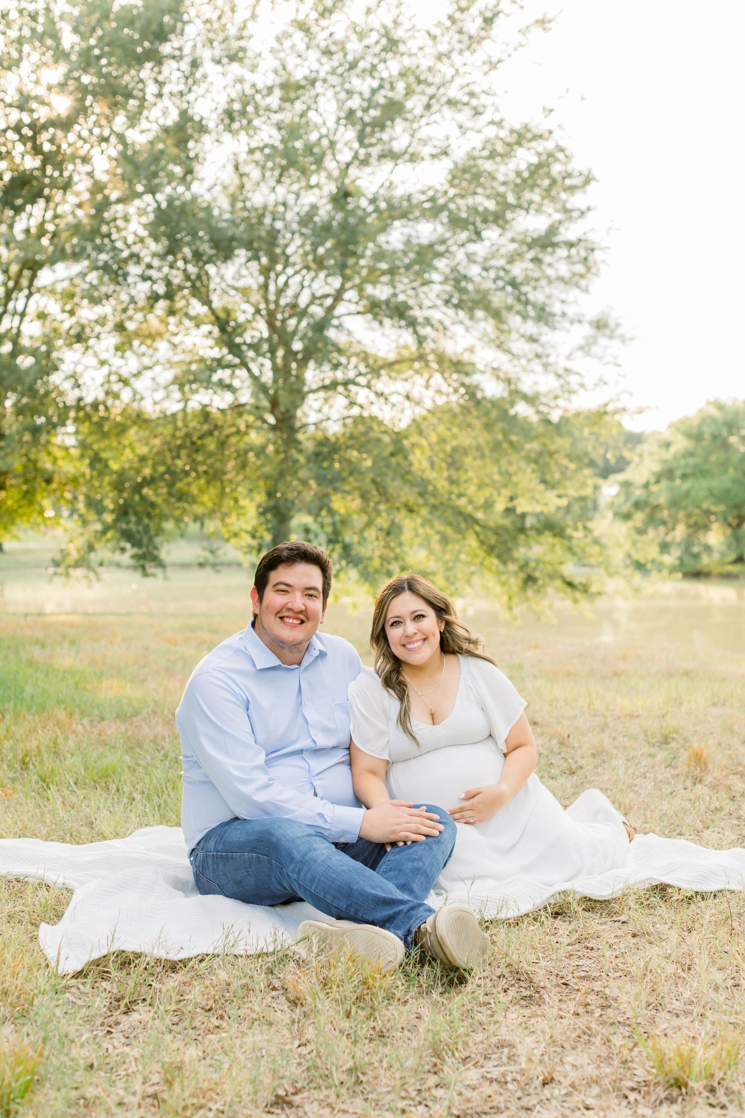 Couple sitting on white blanket in sunny field near pond.