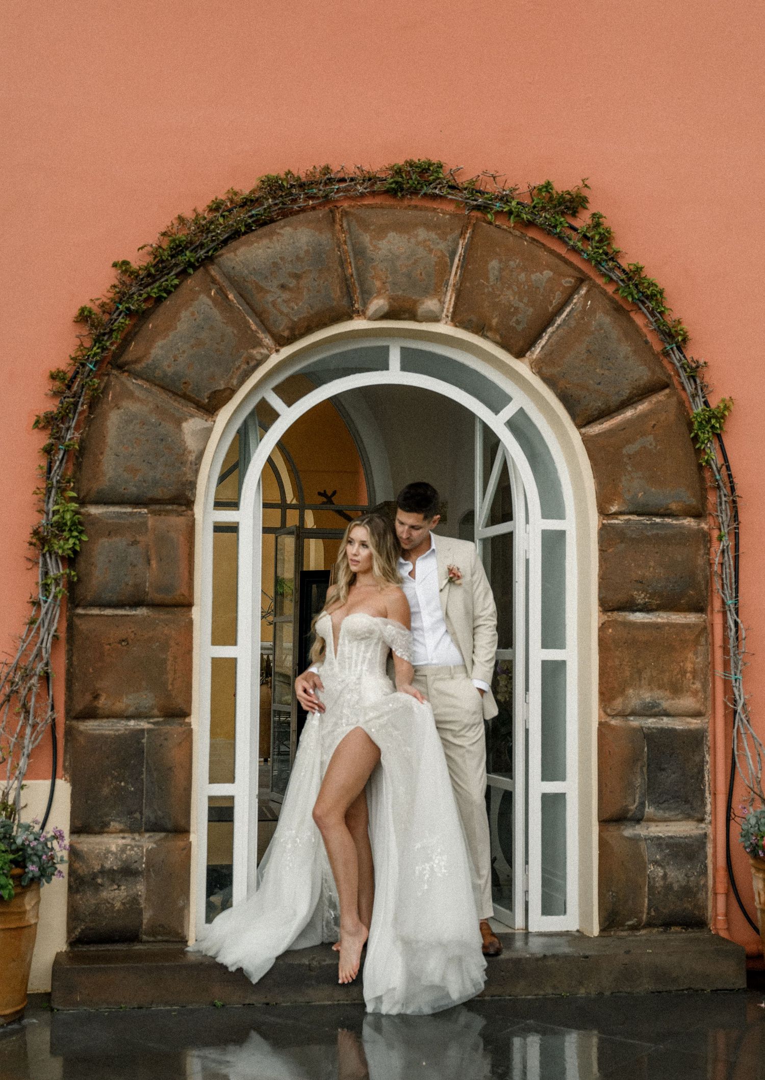 A couple poses in an elegant arched doorway decorated with greenery while wearing white wedding attire.