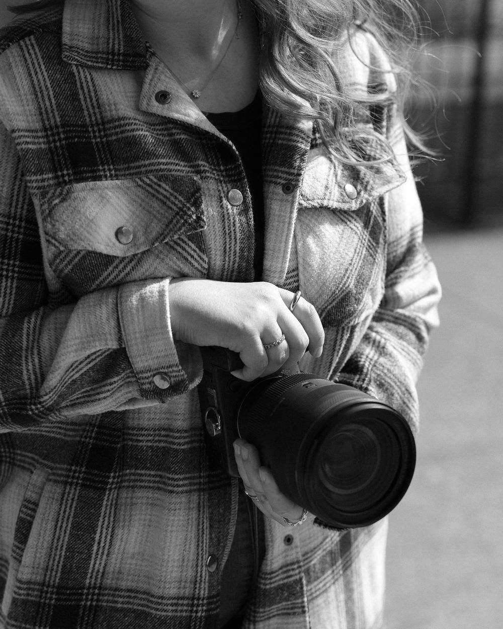 Black and white photo of woman in plaid flannel shirt holding professional camera lens outdoors.