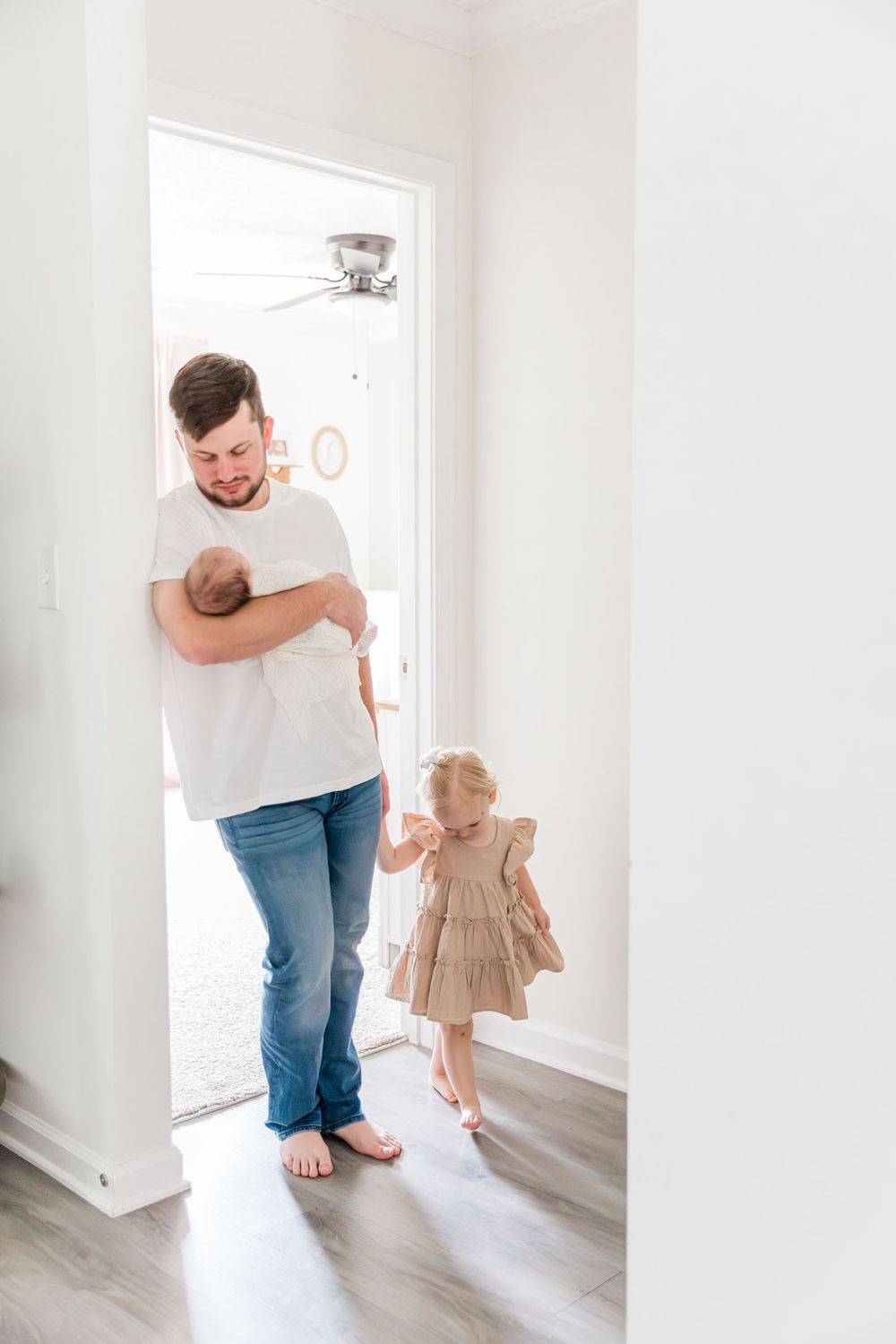 Father holding newborn baby while young toddler girl in tan dress holds his hand in bright white home.