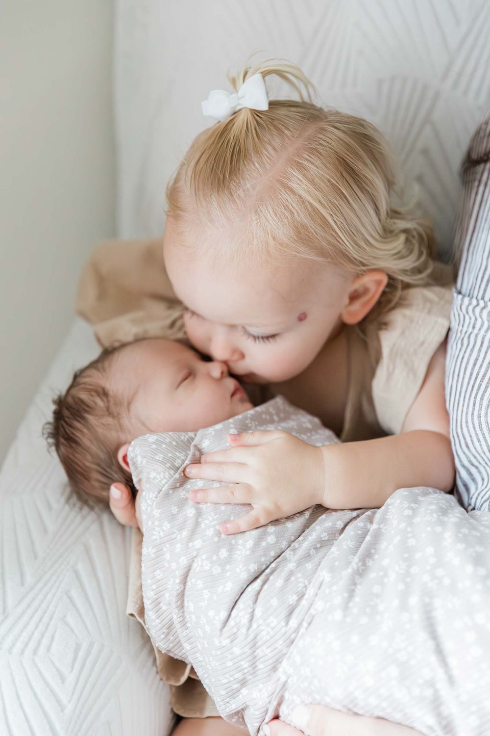 Blonde toddler girl with white bow gently kissing newborn baby sibling wrapped in gray swaddle blanket.