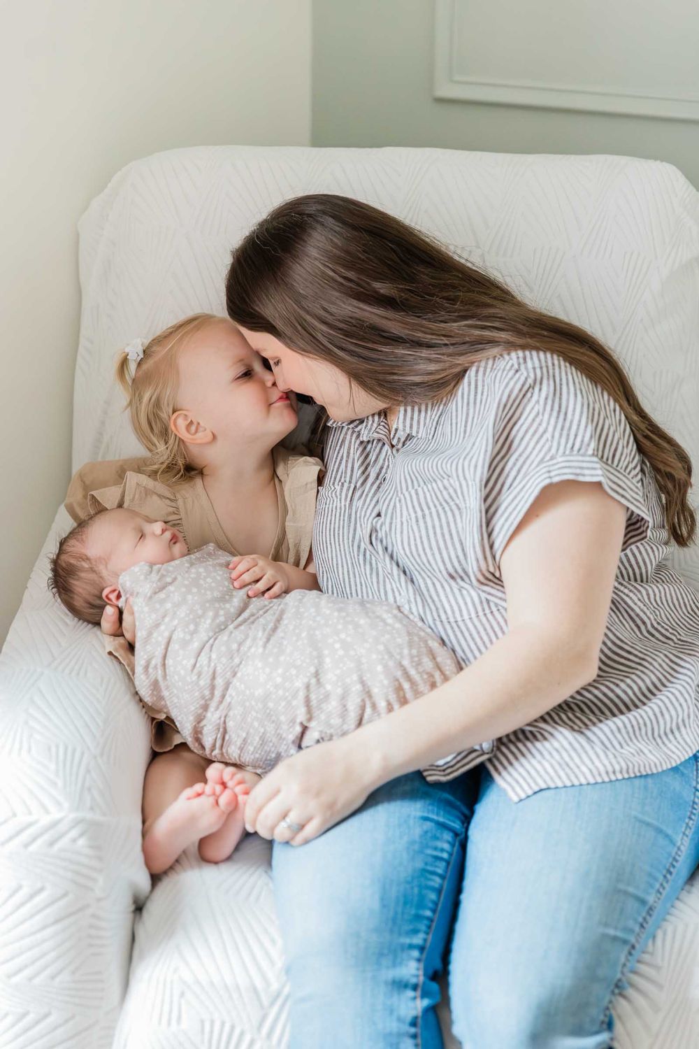 A mother sits in a white chair holding a newborn while sharing a sweet nose kiss with her toddler daughter.