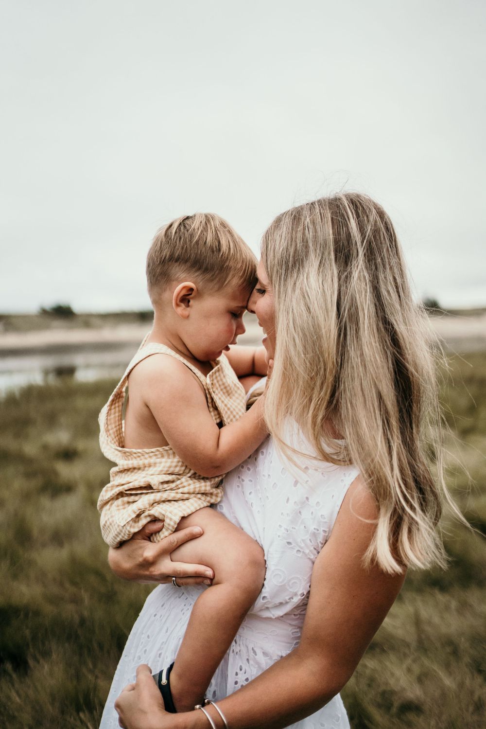 Intimate mother and child moment in white dress against coastal backdrop.