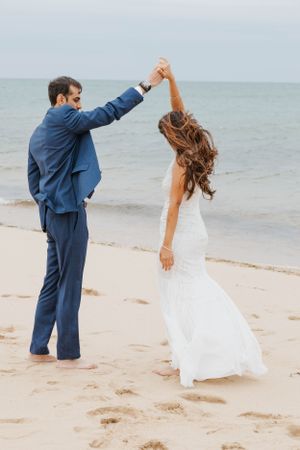Groom in blue suit twirls bride in white wedding gown on a sandy beach by the ocean.