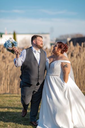 Bride and groom walking through golden reeds outdoors, bride in off-shoulder ball gown holding blue hydrangea bouquet.