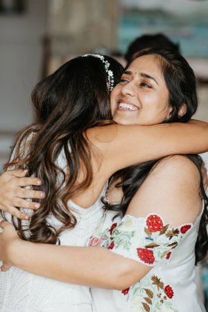 Two women share a warm embrace at a wedding celebration, one wearing a floral embroidered off-shoulder dress.