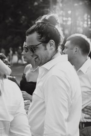 Black and white photo of smiling man with bun hairstyle and sunglasses at outdoor event.