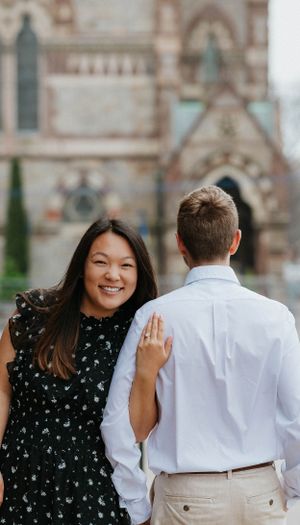 Smiling bride-to-be in black floral dress showing engagement ring while walking with partner near gothic church.
