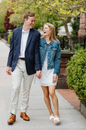 Happy couple strolling and laughing on sunny city sidewalk surrounded by spring greenery, holding hands.