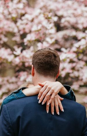 Close-up of woman's hand with engagement ring resting on man's shoulder in front of pink cherry blossoms.