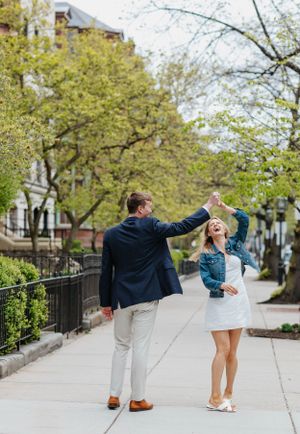 Joyful couple dancing on urban sidewalk in spring, man in navy blazer spinning woman in white dress and denim jacket.