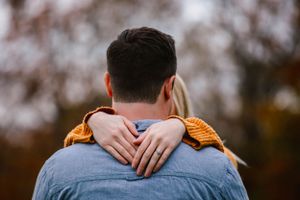Romantic couple embracing from behind with woman's hand on man's neck in autumn setting.