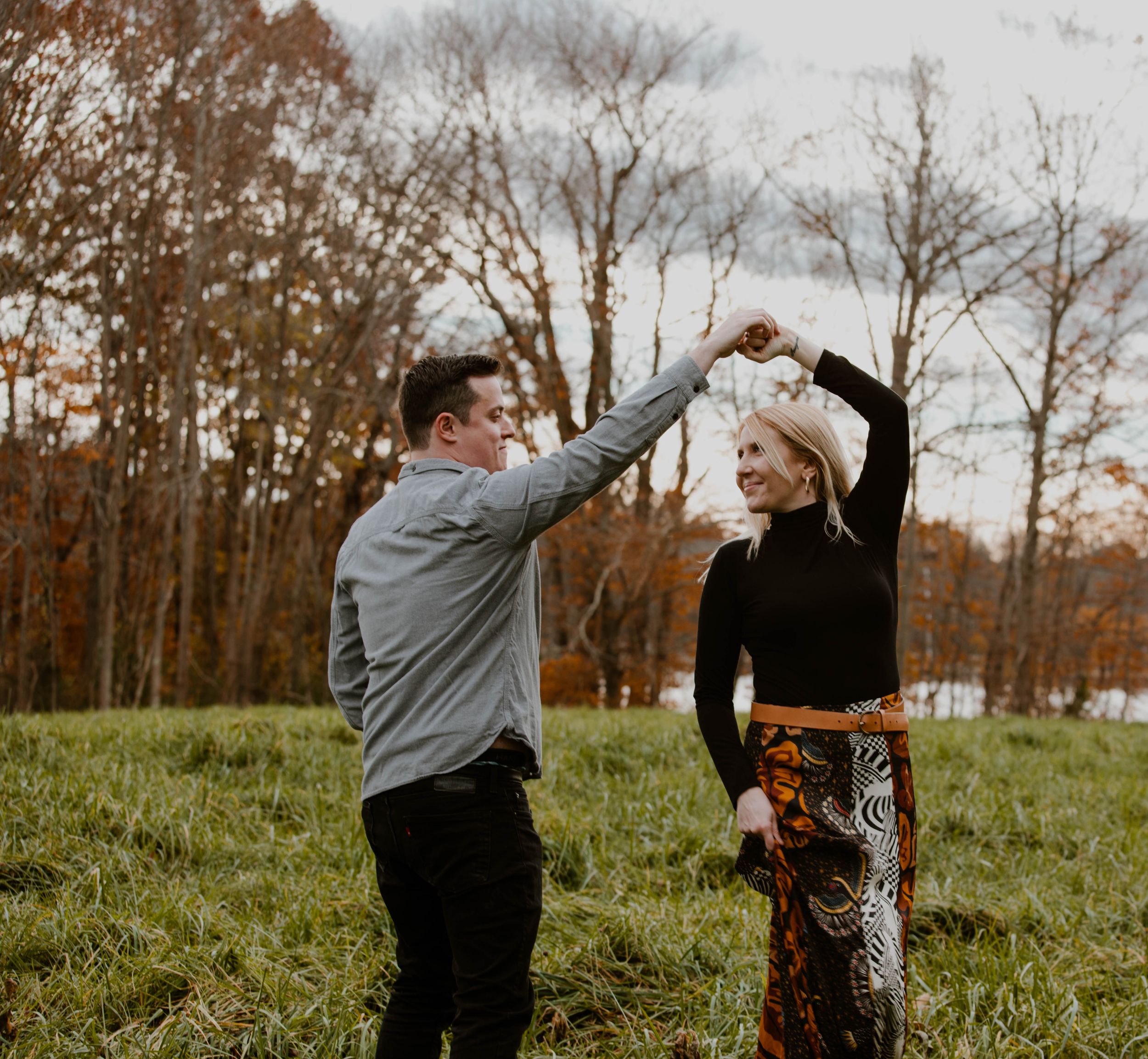 Joyful couple dancing in open field surrounded by colorful autumn trees during fall engagement session.