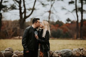 Couple sharing a kiss near stone wall surrounded by fall foliage during engagement photos.