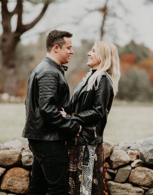 Smiling couple in matching black leather jackets facing each other near rustic stone wall.